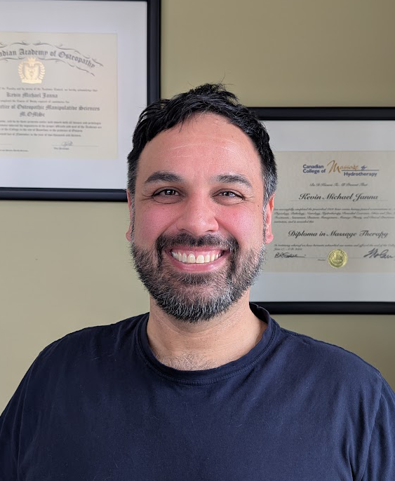 Kevin Janna smiling in a navy t-shirt, with his osteopathy and massage therapy diplomas framed on the wall behind him.
